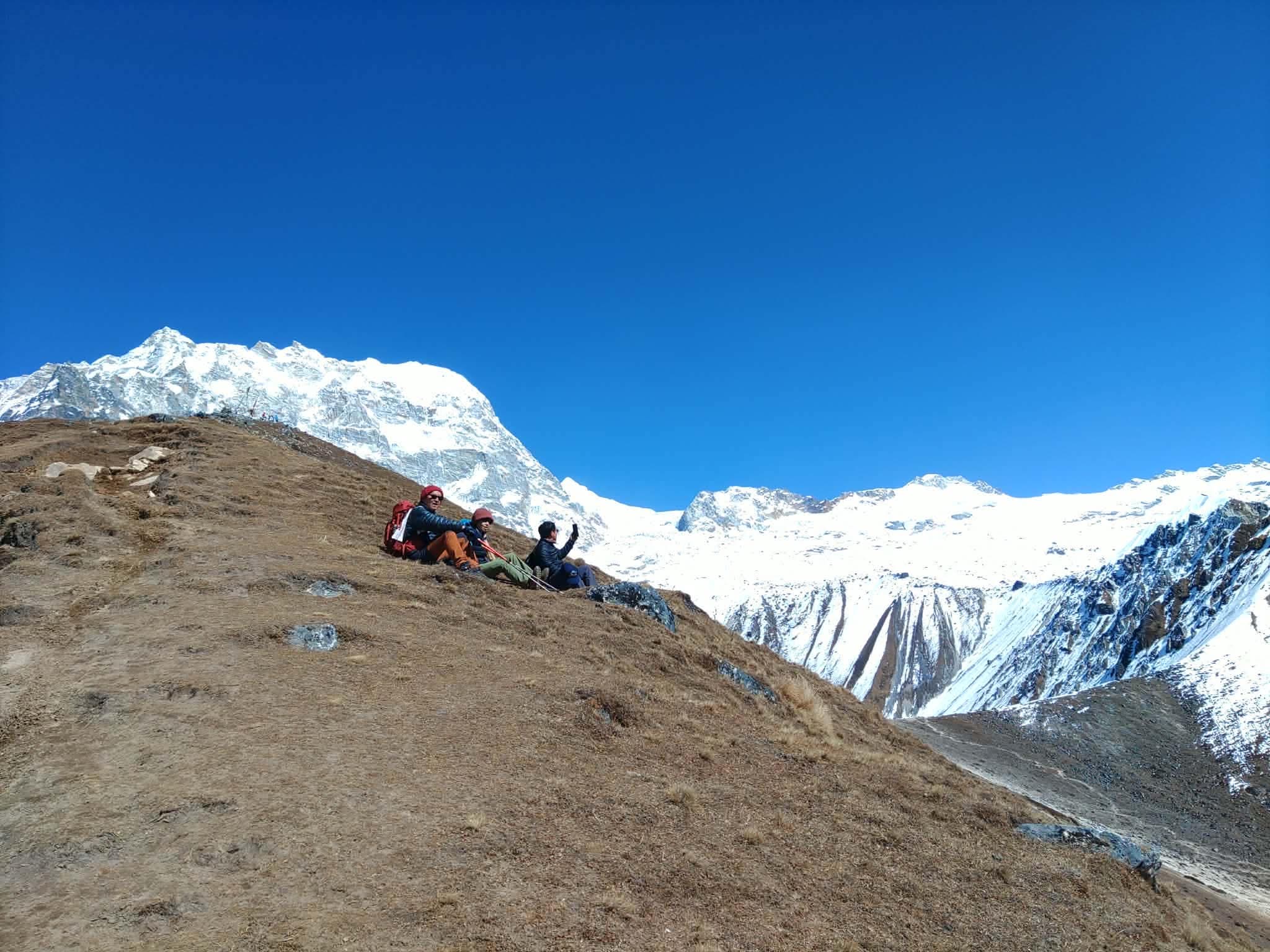 Walkers resting on an open hillside with a snow-covered range behind them.